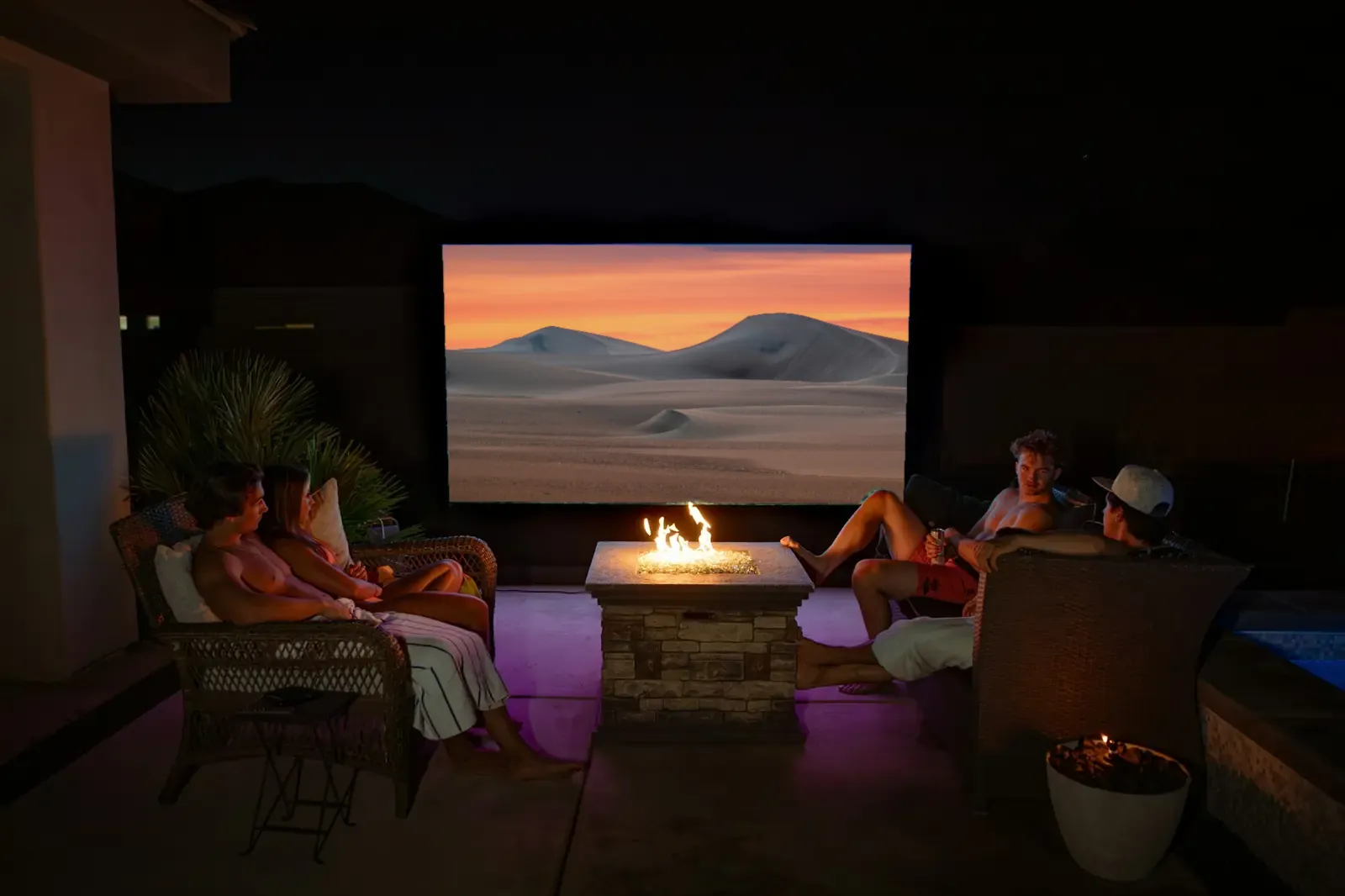 Amigos alrededor de una fogata viendo una pantalla grande al aire libre de noche